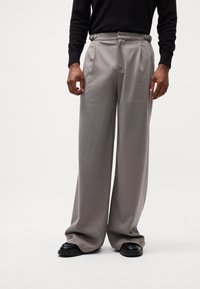 Pantalons larges gris avec plis à l'avant, taille ceinturée et tissu lisse. Associés à des chaussures noires, montrant un design moderne et bien ajusté.