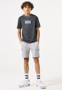 T-shirt in cotone grigio con stampa grafica, abbinato a pantaloni cargo grigi chiari. Indossato con sneakers bianche e calzini a righe bianchi e neri.