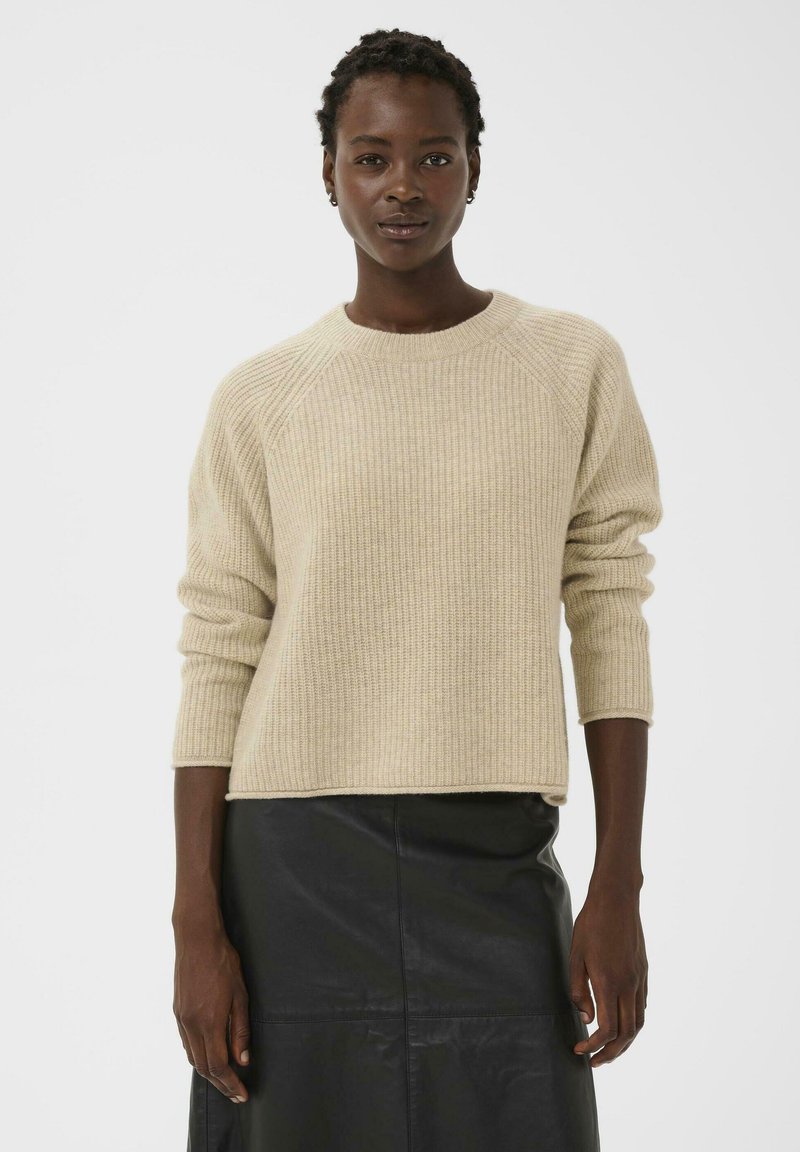 Pull en maille côtelée beige avec un col rond et des manches trois-quarts ; associé à une jupe en cuir noire, mettant en valeur une coupe décontractée.