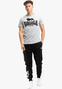 Pilkas medvilninis marškinėliai su juodu liūto logotipu ir tekstu "Lonsdale London", derinami su juodais sportiniais kelnėmis ir baltais sportiniais bateliais.