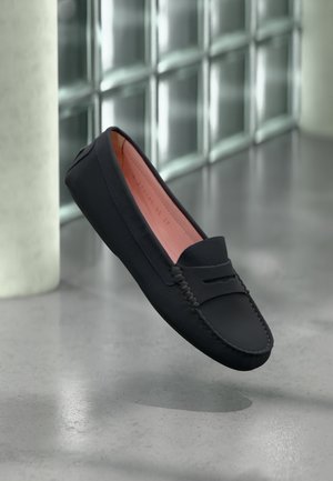 Czarny skórzany mokasyn typu penny loafer z widocznym przeszyciem, różowym wnętrzem, unoszący się nad pokrytą polerowanym betonem podłogą w pobliżu ściany z bloków szklanych.