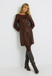 Robe en sequins marron avec des manches longues et un col bateau ; longueur genou avec une coupe ample. Associée à des bottes à talons marron montantes.