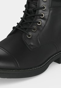 Bottine en cuir noire à lacets avec œillets métalliques et semelle en caoutchouc cousue sur fond blanc.