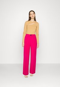Top beige a maniche lunghe con dettaglio a taglio diagonale abbinato a pantaloni a vita alta e gamba larga di un rosa brillante. Sandali semplici completano l'outfit.
