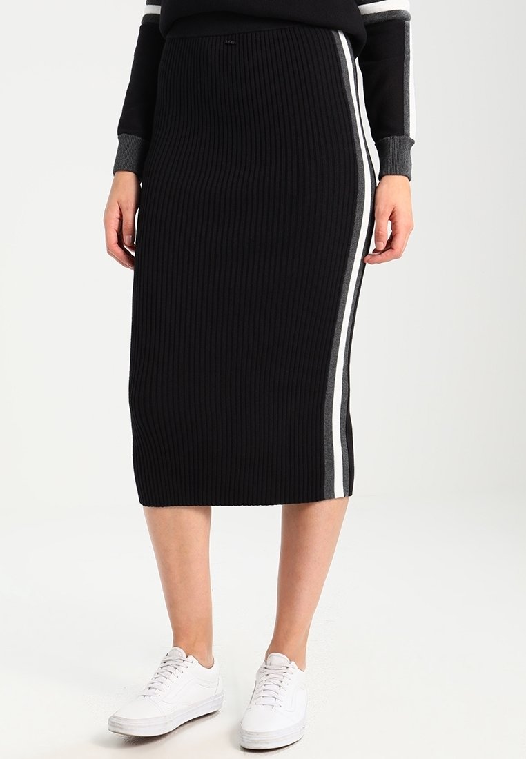 Femme portant une jupe midi côtelée noire avec des bandes latérales blanches et grises, associée à des baskets blanches et un haut noir.