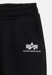 Black sweatpants with an elastic waistband, side pocket, and a prominent white logo reading "ALPHA INDUSTRIES" with three horizontal lines.