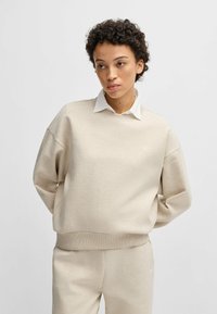 Felpa beige con vestibilità rilassata, polsini e orlo a costine. Camicia bianca con colletto sotto, con un piccolo logo sul petto.
