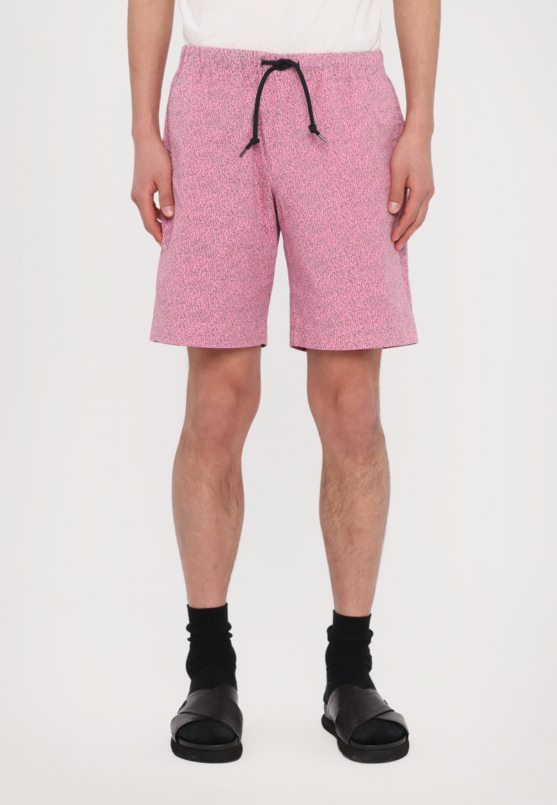 Roze zwembroek met een gestructureerd patroon, elastische tailleband en zwarte trekkoord. Draagt zwarte sokken en zwarte sandalen.