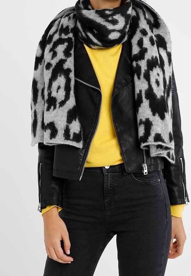 Femme portant une écharpe à motif léopard noir et blanc, une veste en cuir noire, un pull jaune et un jean noir, debout les bras détendus.