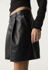 Shorts en cuir noir à texture lisse, dotés de plis à l'avant et d'une ceinture avec fermeture par bouton. Mis en valeur par un haut transparent orné de petits embellissements.