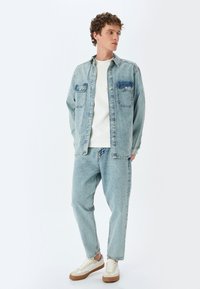 Giacca di jeans azzurro chiaro con due tasche frontali, maglietta bianca a girocollo e jeans larghi coordinati. Le sneakers presentano dettagli beige.