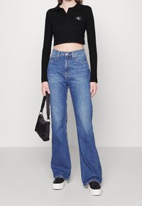 Kvinna som bär en svart croppad långärmad polo, högmidjade lösa blå jeans, svartvita sneakers och håller en liten svart handväska.