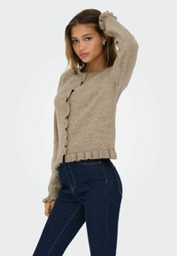 Gai bēšas krāsas kardigāns ar viļņotām malām un pogām, izgatavots no mīksta dzijas materiāla; saskaņots ar tumši zilām augsto jostasvietu džinsām.