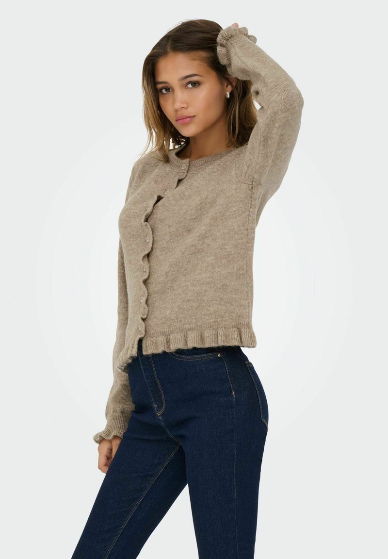 Gai bēšas krāsas kardigāns ar viļņotām malām un pogām, izgatavots no mīksta dzijas materiāla; saskaņots ar tumši zilām augsto jostasvietu džinsām.