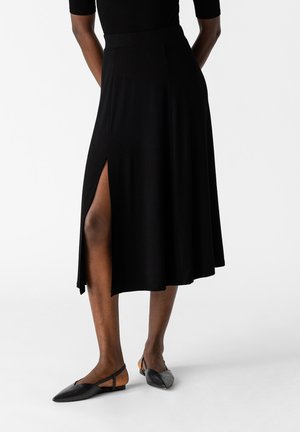Falda negra en corte A con una abertura lateral, hecha de un tejido suave. Combinada con unos zapatos planos negros con punta, que cuentan con un diseño elegante y detalles de correa.