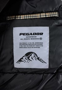 L'etichetta di un capo d'abbigliamento nero presenta il logo "PEGADOR", su uno sfondo bianco e una grafica montana. Striscia a quadri sopra l'etichetta.