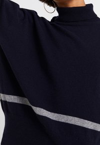 Frau trägt einen dunkelblauen Rollkragenpullover mit einem hellgrauen horizontalen Streifen und großen goldenen Creolen.