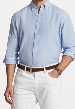 Mężczyzna w jasno-niebieskiej koszuli na guziki z podwiniętymi rękawami, białych spodniach, brązowym skórzanym pasku i brązowym zegarku z metalową tarczą.