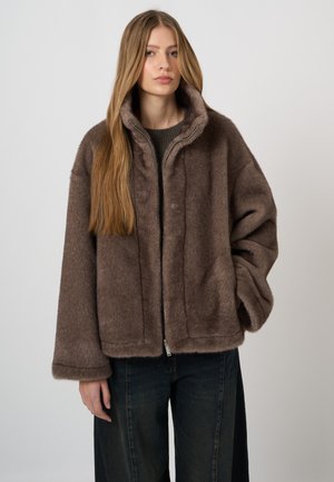 Junge Frau mit langen, glatten Haaren, die eine braune, flauschige Jacke mit Reißverschluss und dunkle, weite Hosen trägt, vor einem einfarbigen Hintergrund.