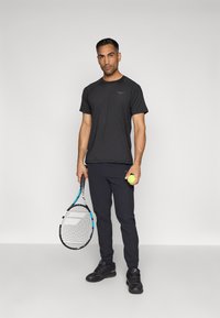 Černé sportovní tričko s krátkými rukávy, spárované s tmavými sportovními kalhotami. Model drží zelený tenisový míček a raketu s modrými akcenty.