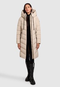 Vrouw in een lange beige pufferjas met grote capuchon, zwarte legging en zwarte laarzen tot de knie, staand op een effen lichte achtergrond.