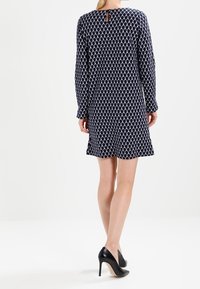 Marineblaues Kleid mit geometrischem weißen Muster, langen Ärmeln, Rundhalsausschnitt und Rückenschlitz-Detail. Kombiniert mit schwarzen High Heels.