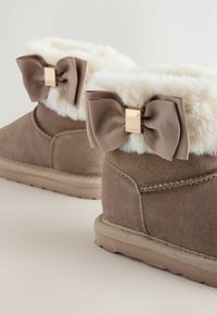Bruine suède enkelbootjes met een crèmekleurige nepbont voering, voorzien van twee textuurstrikken en vierkante gouden hardware accenten aan de buitenzijde.