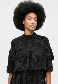 Kvinna med naturlig afrofrisyr klädd i en svart prickig höghalserock, som står mot en ljus neutral bakgrund.