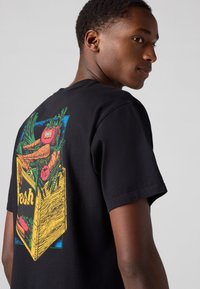 Black cotton t-shirt featuring a colorful vegetable crate graphic on the back, with bold colors and detailed patterns. Short sleeves.