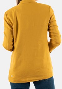 Marynarka w kolorze musztardowej żółci o dopasowanym kroju, z długimi rękawami, klapami z wycięciem oraz gładką fakturą. Posiada pojedynczy rozcięcie z tyłu i subtelne logo.
