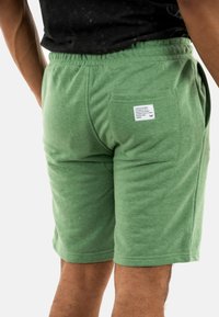 Pantalones cortos de algodón verde con cintura elástica, bolsillos laterales y un bolsillo trasero de parche que presenta una pequeña etiqueta de marca. Diseño suave y casual.