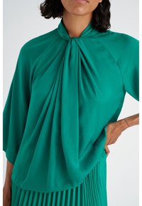 Blusa teal con parte anteriore drappeggiata, dettagli plissettati e colletto alto. Tessuto morbido e liscio con maniche a tre quarti. Design semplice ed elegante.