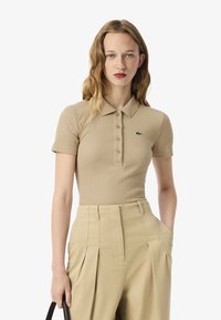 Polo shirt beige a coste con colletto e patta a cinque bottoni, caratterizzato dal logo verde Lacoste, abbinato a pantaloni beige a vita alta.