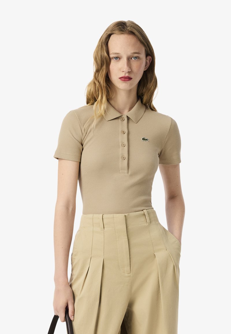 Polo shirt beige a coste con colletto e patta a cinque bottoni, caratterizzato dal logo verde Lacoste, abbinato a pantaloni beige a vita alta.