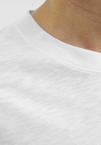 Hvid bomulds T-shirt med en struktureret overflade, med rund hals og flade sømme, vist delvist over en hudfarvet baggrund.