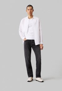 Camicia di lino bianca sopra una canotta bianca, abbinata a jeans di denim scuri e sneakers chiare. Design semplice, taglio dritto, look casual.