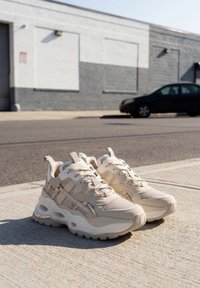 Par beige chunky sneakers placeret på en solbelyst fortov med en sort bil og en grå bygning i baggrunden.