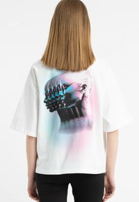 Biała, oversize t-shirt z grafiką przedstawiającą wiele głów w odcieniach niebieskiego i różowego na plecach. Krótkie rękawy, miękki bawełniany materiał.