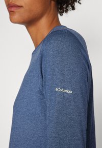 Osoba v melírovaném modrém tričku s dlouhým rukávem a logem "Columbia" na horní části rukávu, zobrazená z boku na jednoduchém pozadí.