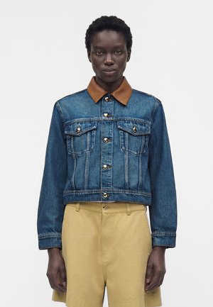 Donkerhuidig persoon die een blauwe spijkerjas draagt met een bruine kraag en beige short met hoge taille, staand tegen een effen witte achtergrond.