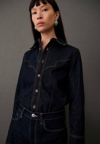 Dunkelblauses Denimhemd mit einem Yoke-Design im Western-Stil, ausgestattet mit einer durchgehenden Knopfleiste und silbernen Druckknöpfen. Getragen mit dunkelblauen Denimhosen.