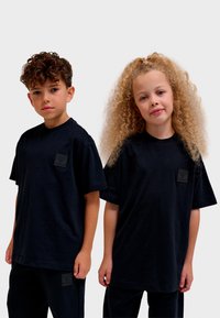 Svarta t-shirts i bomull med korta ärmar och en fyrkantig logotyp på bröstet, båda bärs av två barn i avslappnade poser.