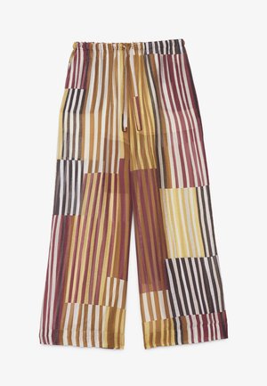 Vrouw met kort donker haar draagt een los shirt en broek met verticale meerkleurige strepen in bruin, geel en bordeaux, staande op een grijze vloer.