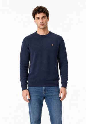 Giovane uomo con capelli ricci indossa un maglione blu navy girocollo e jeans blu su uno sfondo bianco semplice.