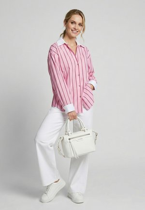 Žena v růžovém pruhovaném tričku, bílých kalhotách a teniskách držící bílou kabelku, usmívající se na jednobarevném světle pozadí.