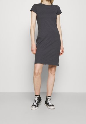 Kobieta w dopasowanej, krótkim rękawem sukience w kolorze węgla, zestawiona z czarnymi sneakersami za kostkę, stoi na jednolitym jasnym tle.