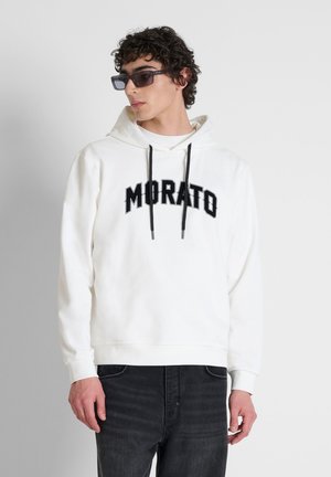 Giovane uomo che indossa occhiali da sole neri, una felpa bianca con cappuccio con il testo "MORATO" e jeans neri, che guarda di lato su uno sfondo semplice.