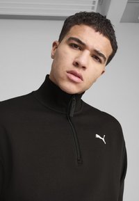 Sort zip-up pullover lavet af ribbet stof, med høj krave og et lille hvidt Puma-logo på venstre bryst.