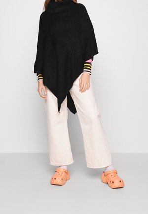Personne portant un poncho asymétrique noir, une chemise rayée à manches longues, un pantalon blanc large, et des sabots orange avec des chaussettes blanches sur fond uni.