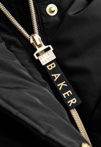 Tirador de cremallera dorado con texto "BYTED BAKER" en tela negra y una lengüeta de tela azul marino con relieve "BAKER" en letras doradas.